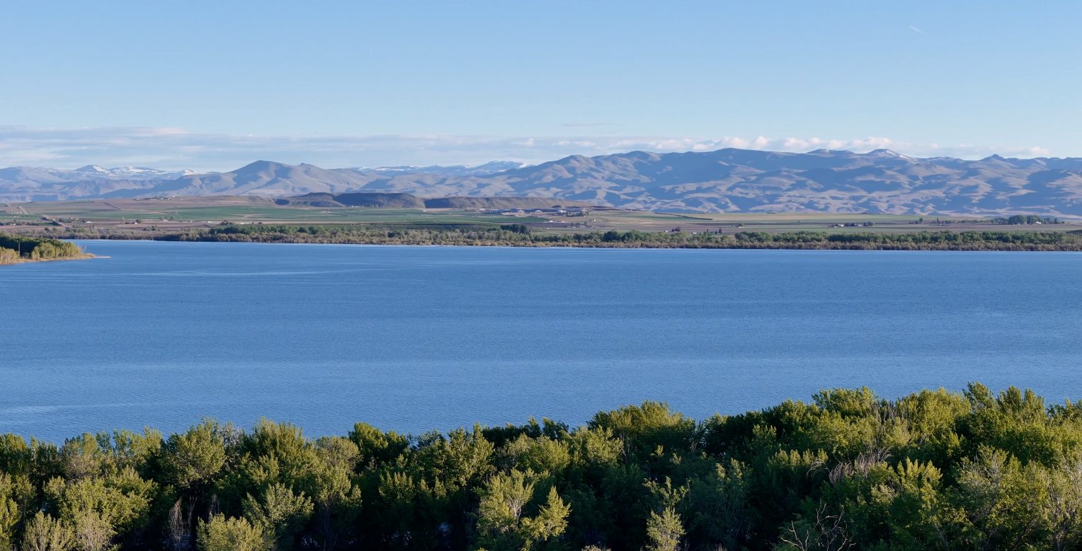 Lake Lowell in Nampa, Idaho with calm blue water, rolling farmland, and the Owyhee Mountains in the distance.